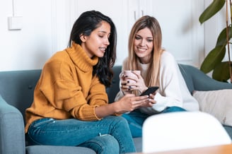 shot-two-pretty-young-women-using-mobile-phone-while-drinking-coffee-sofa-home